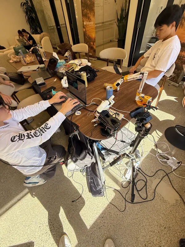 Around 4 pm: the SO-101 at the edge of the table, a camera on a tripod beside it, laptops everywhere